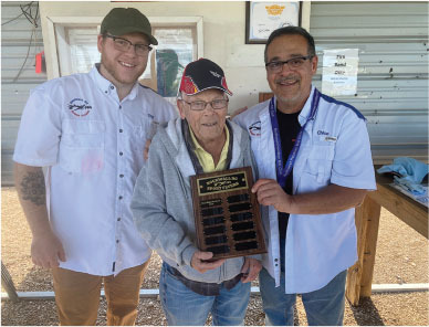 Three men smiling, one holding a plaque, standing outside in casual attire.