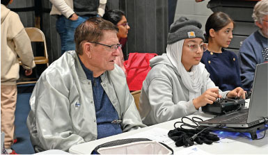 Two people sitting at a table with a laptop, others in the background.