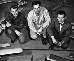 Three men kneeling on a floor with model airplanes around them.