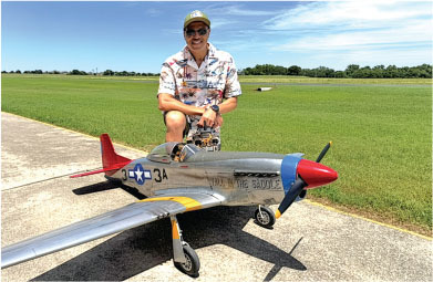 A person smiles kneeling by a large model airplane on a sunny day.