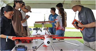 Students examining a red model aircraft on a table under a canopy.