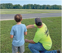 Man and boy facing runway, man pointing, grass and trees in background.
