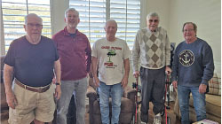 Five elderly men smiling in a living room, one using a walking aid.