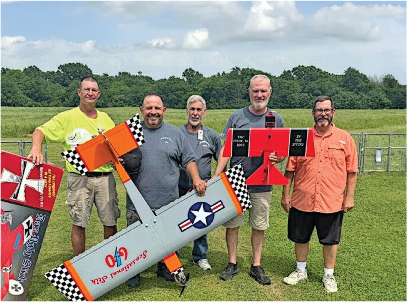 Five men holding model airplanes on a grassy field, smiling at the camera.