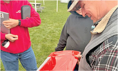 Three people outdoors examining a red tray or object, grass in the background.