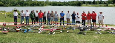 Group of people with remote-control planes by a lake, green grass, and trees.