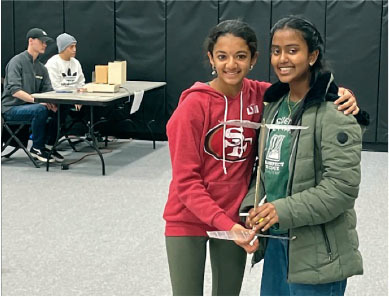 Two girls smiling, holding a trophy in a gym.