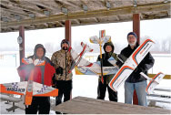 Four people holding model airplanes under a wooden shelter in winter.