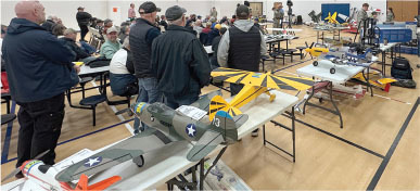 Model airplanes displayed on tables at an indoor event with people gathered around.