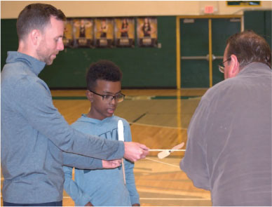 Three people in a gym discussing a small rocket model.
