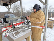 Person in a heavy coat adjusts a model airplane on a snowy table.