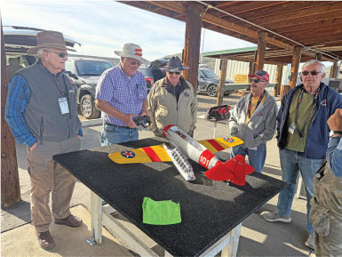 Six people standing around a model airplane on a table outdoors.