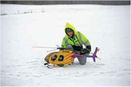 Person in a green jacket kneeling in snow with a large model helicopter.