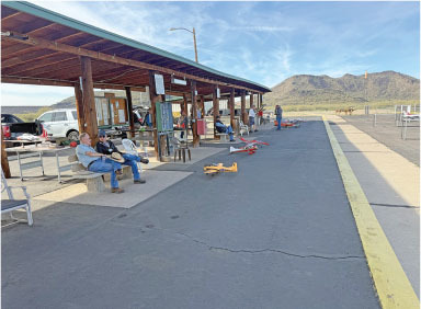 Outdoor pavilion with people sitting, model airplanes on ground, mountains in background.