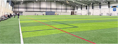 Indoor soccer field with green turf, red and black lines, and people seated on benches.