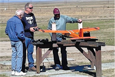 Three men examining an orange model airplane on a stand in a field.