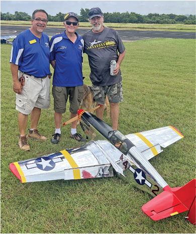 Three men stand on grass, smiling beside a silver model airplane with red and yellow accents.