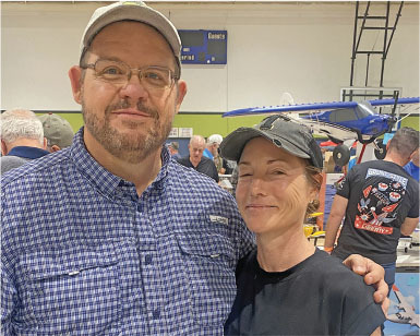 Man and woman smiling at indoor event, a model plane in the background.