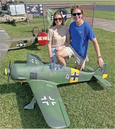 Two people kneeling by a green model warplane on grass.