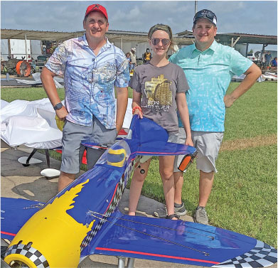 Three people standing behind a blue and yellow model airplane on a sunny day.
