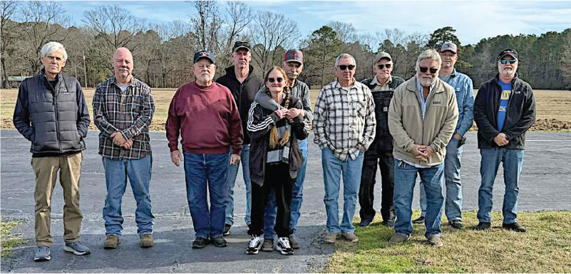 Group of eleven people outdoors on a paved area near a field, wearing casual clothing.