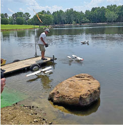 Man operating model seaplanes on a lake dock, surrounded by trees.