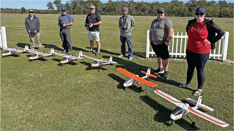 Seven people with remote-controlled planes on a grassy field on a sunny day.