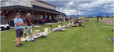 Two men stand by remote-controlled planes on a grassy field, with people in the background.