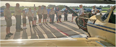 Group of people in uniform listening to a person near a small aircraft on a sunny day.