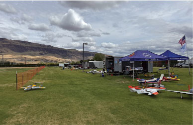 Model airplanes displayed on grass, under tents, with mountains and cloudy sky in background.