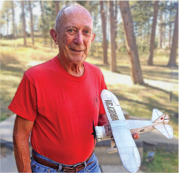 Elderly man in a red shirt holding a model airplane outside.