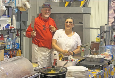 Two people in a kitchen, one holding a spatula, smiling at the camera.