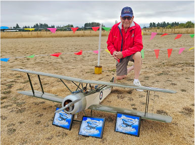 Man kneeling beside large model biplane with plaques on dry grass, colorful flags in background.