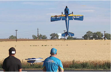 Two men watch a blue model airplane in the sky above a field.