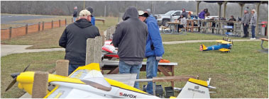 People preparing model airplanes outdoors, with tables and grass field.