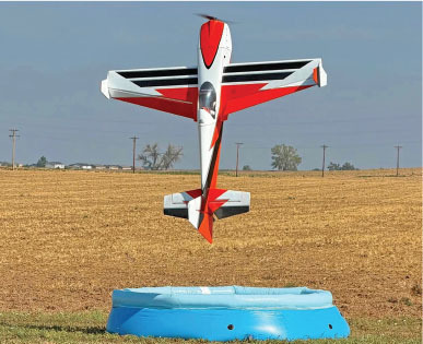 Red and white model plane hovering above a small blue pool in a field.