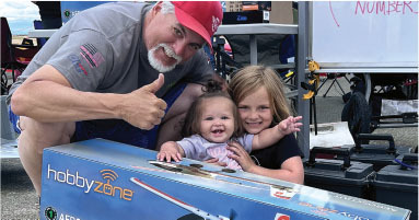 Man with two children and large HobbyZone box, outdoors, daytime.