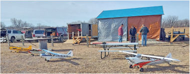 Model airplanes on the ground, people and vehicles beside a shed.