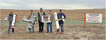 Five people holding model airplanes next to a field sign, cloudy sky above.