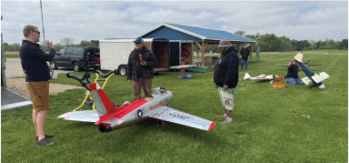 Three men with scale model planes on grass, blue shed in background.