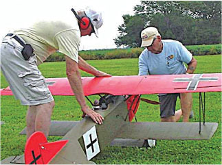 Two men adjusting a large, red model airplane on grass.