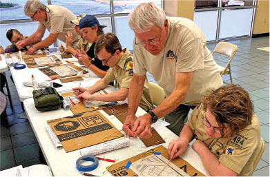 Scouts and leaders work on cardboard crafts at a table indoors.