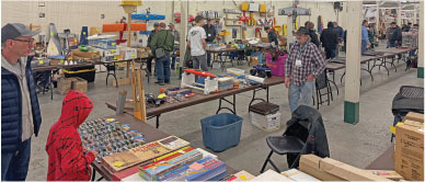 A hobby show with people browsing tables of model airplanes and related items.