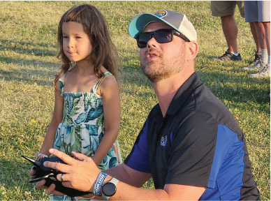 Man teaching girl to fly drone in park, sunny day.