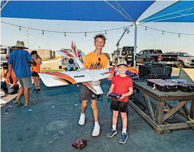 Two smiling boys holding a model airplane and remote, under a blue canopy.