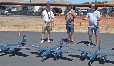 Three men standing behind model planes on an airfield, giving thumbs up.
