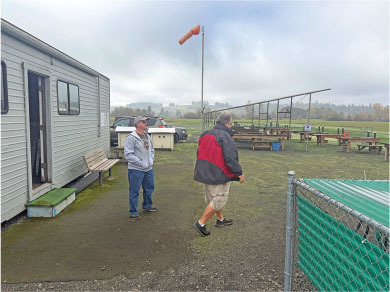 Two men near a building, wind sock in background, cloudy sky.