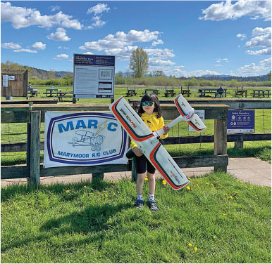 Child holding model plane, standing by Marymoor R/C Club sign on a sunny day.