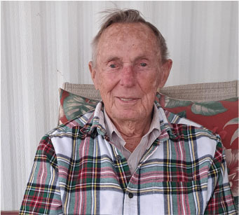Elderly man in plaid shirt smiling, seated against a floral pillow.