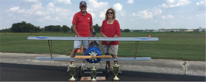 Two people in red shirts stand behind a model biplane with trophies on the ground.